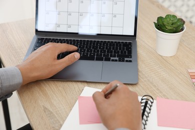 Photo of Man writing in business planner and using laptop at wooden table in office, closeup