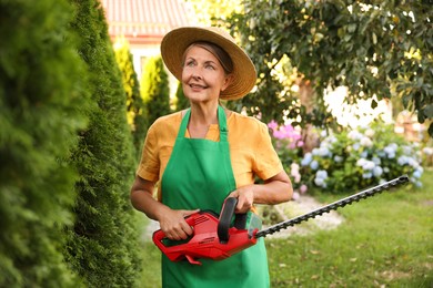 Photo of Smiling senior woman in hat and apron with electrical hedge trimmer in garden