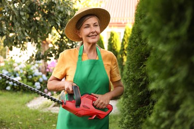 Photo of Smiling senior woman in hat and apron with electrical hedge trimmer in garden