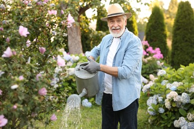 Photo of Elderly man in hat watering beautiful flowers with can in garden
