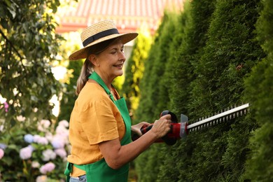 Photo of Smiling senior woman in hat and apron working with electrical hedge trimmer in garden