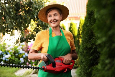 Photo of Smiling senior woman in hat and apron with electrical hedge trimmer in garden