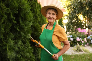 Photo of Senior woman in hat and apron with manual hedge trimmer working in garden