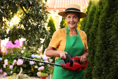 Photo of Smiling senior woman in hat and apron with electrical hedge trimmer in garden