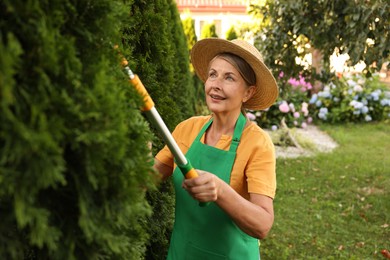 Photo of Senior woman in hat and apron with manual hedge trimmer working in garden