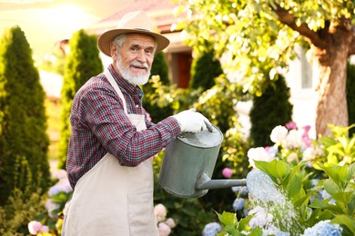 Photo of Elderly man in hat and apron watering beautiful flowers with can in garden