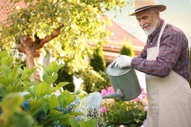 Photo of Elderly man in hat and apron watering beautiful flowers with can in garden