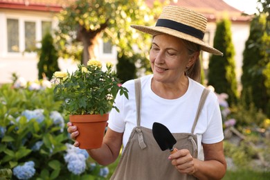 Photo of Smiling senior woman in hat and apron with potted flowers and shovel in garden