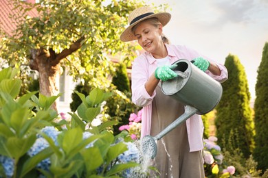 Photo of Smiling senior woman in hat and apron watering beautiful flowers with can in garden