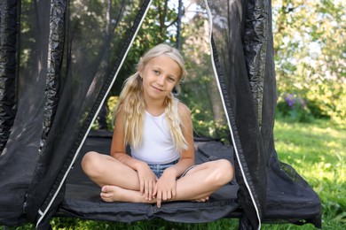 Photo of Cute little girl sitting on black trampoline outdoors