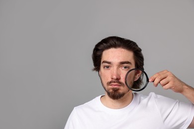 Photo of Young man with acne problem holding magnifying glass near his face on light grey background. Space for text