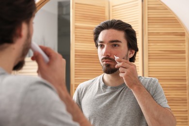 Photo of Young man with acne problem applying cream onto his face near mirror at home