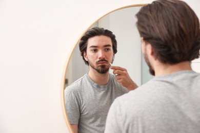 Photo of Young man with acne problem applying cream onto his face near mirror at home, space for text
