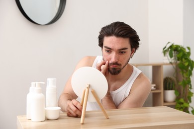 Photo of Young man with acne problem looking at mirror at table indoors