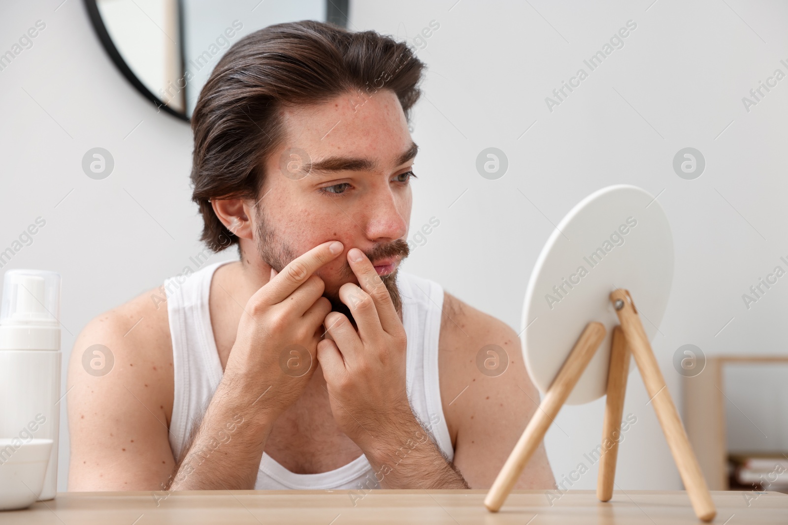 Young man with acne problem looking at mirror at table indoors Photo of Young man with acne problem looking at mirror at table indoors