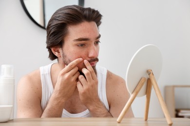 Photo of Young man with acne problem looking at mirror at table indoors