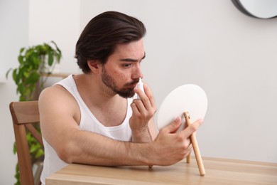 Photo of Young man with acne problem applying cream onto his face near mirror at table indoors