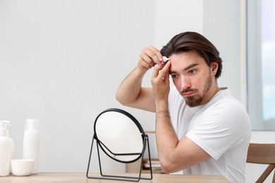 Photo of Young man with acne problem applying cream onto his face near mirror at table indoors