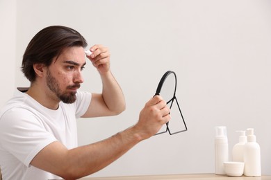 Photo of Young man with acne problem applying cream onto his face near mirror at table indoors