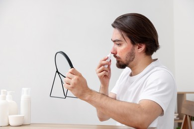 Photo of Young man with acne problem applying cream onto his face near mirror at table indoors