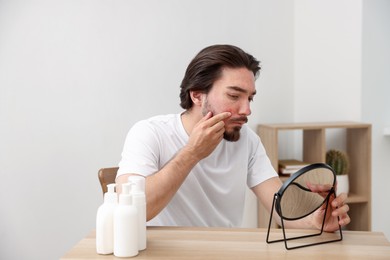 Photo of Young man with acne problem looking at mirror at table indoors