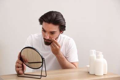 Photo of Young man with acne problem looking at mirror at table indoors