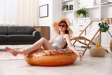 Photo of Smiling woman in sunglasses and straw hat with inflatable ring having fun on floor at home. Dreaming about vacation at sea