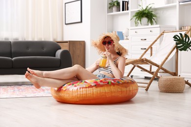 Photo of Woman in sunglasses and straw hat drinking juice from mason jar with inflatable ring on floor at home. Dreaming about vacation at sea