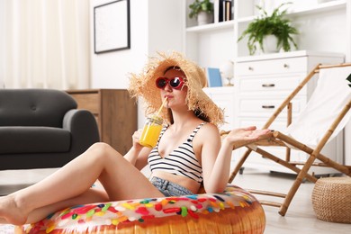 Photo of Woman in sunglasses and straw hat drinking juice from mason jar with inflatable ring on floor at home. Dreaming about vacation at sea