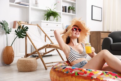 Photo of Happy woman in sunglasses and straw hat with mason jar of juice and inflatable ring having fun on floor at home. Dreaming about vacation at sea