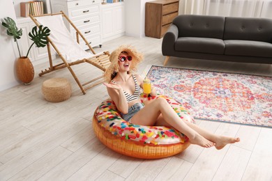 Photo of Happy woman in sunglasses and straw hat with mason jar of juice and inflatable ring having fun on floor at home. Dreaming about vacation at sea