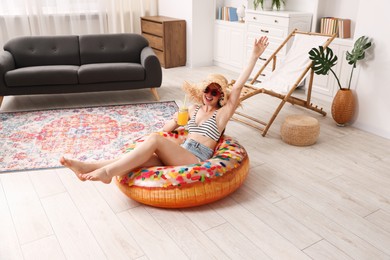 Photo of Happy woman in sunglasses and straw hat with mason jar of juice and inflatable ring having fun on floor at home. Dreaming about vacation at sea
