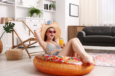 Photo of Happy woman in sunglasses and straw hat with mason jar of juice and inflatable ring having fun on floor at home. Dreaming about vacation at sea