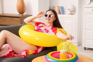 Photo of Happy woman in sunglasses with mason jar of juice and inflatable ring on sofa at home. Dreaming about vacation at sea