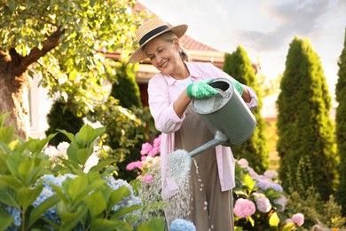 Photo of Smiling senior woman in hat and apron watering beautiful flowers with can in garden