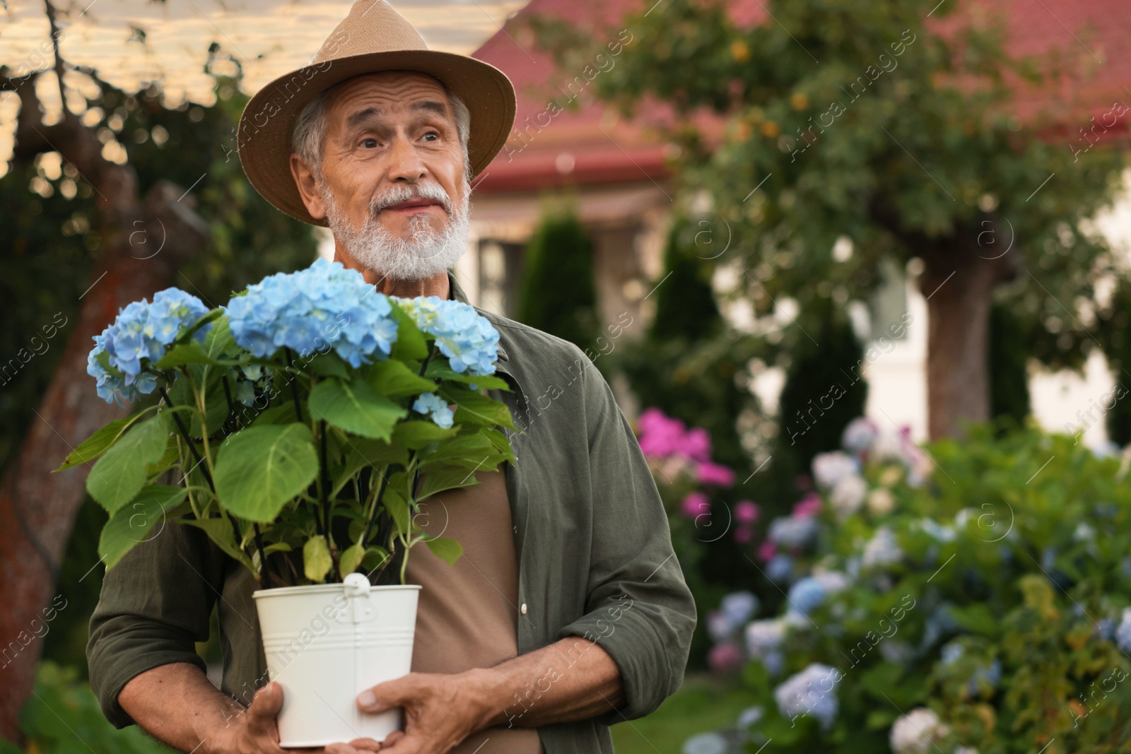 Photo of Elderly man in hat with potted flowers in garden. Space for text