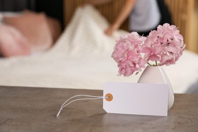 Photo of Welcome. Chambermaid making bed in hotel room, focus on blank card and hydrangea flowers