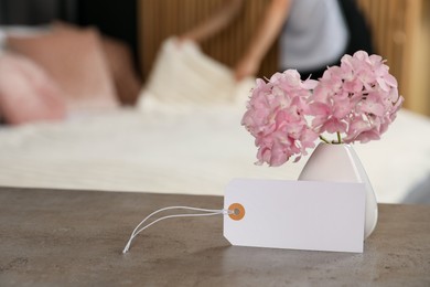 Photo of Welcome. Chambermaid making bed in hotel room, focus on blank card and hydrangea flowers