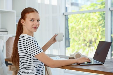 Photo of Teenage girl with cup of drink using laptop at wooden desk indoors. Home workplace