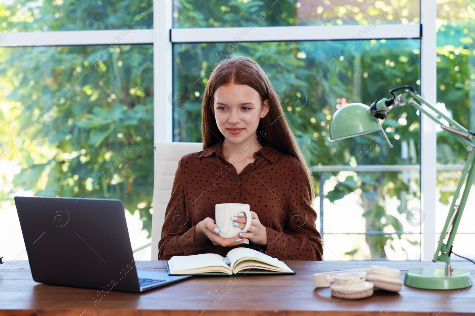 Photo of Teenage girl with cup of drink using laptop at wooden desk indoors. Home workplace