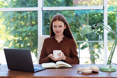 Photo of Teenage girl with cup of drink using laptop at wooden desk indoors. Home workplace