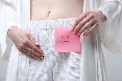 Photo of Woman holding sticky note with drawn sad face and suffering from cystitis symptoms on light grey background, closeup