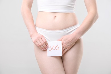 Photo of Woman holding sticky note with word SOS and suffering from cystitis symptoms on light grey background, closeup