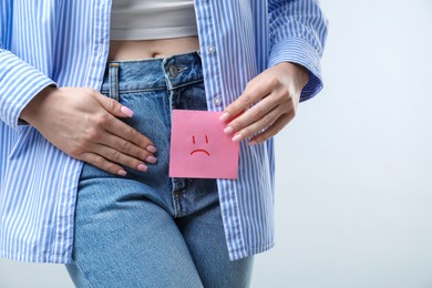 Photo of Woman holding sticky note with drawn sad face and suffering from cystitis symptoms on light grey background, closeup. Space for text