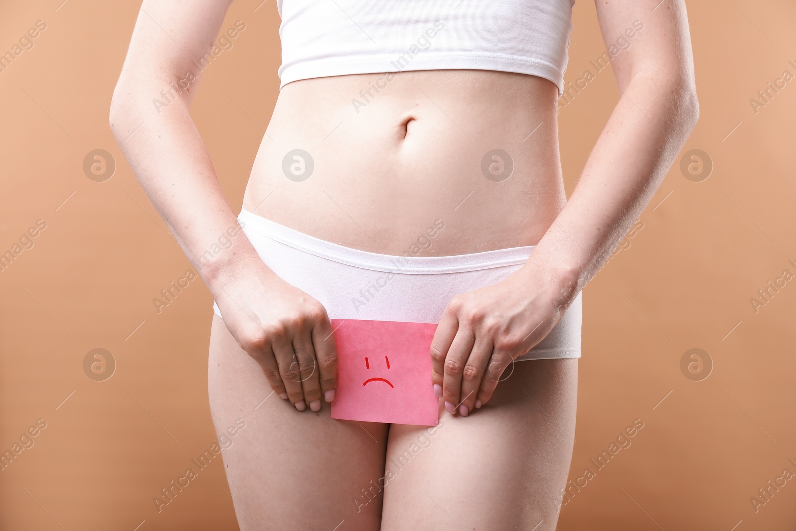 Photo of Woman holding sticky note with drawn sad face and suffering from cystitis symptoms on dark beige background, closeup