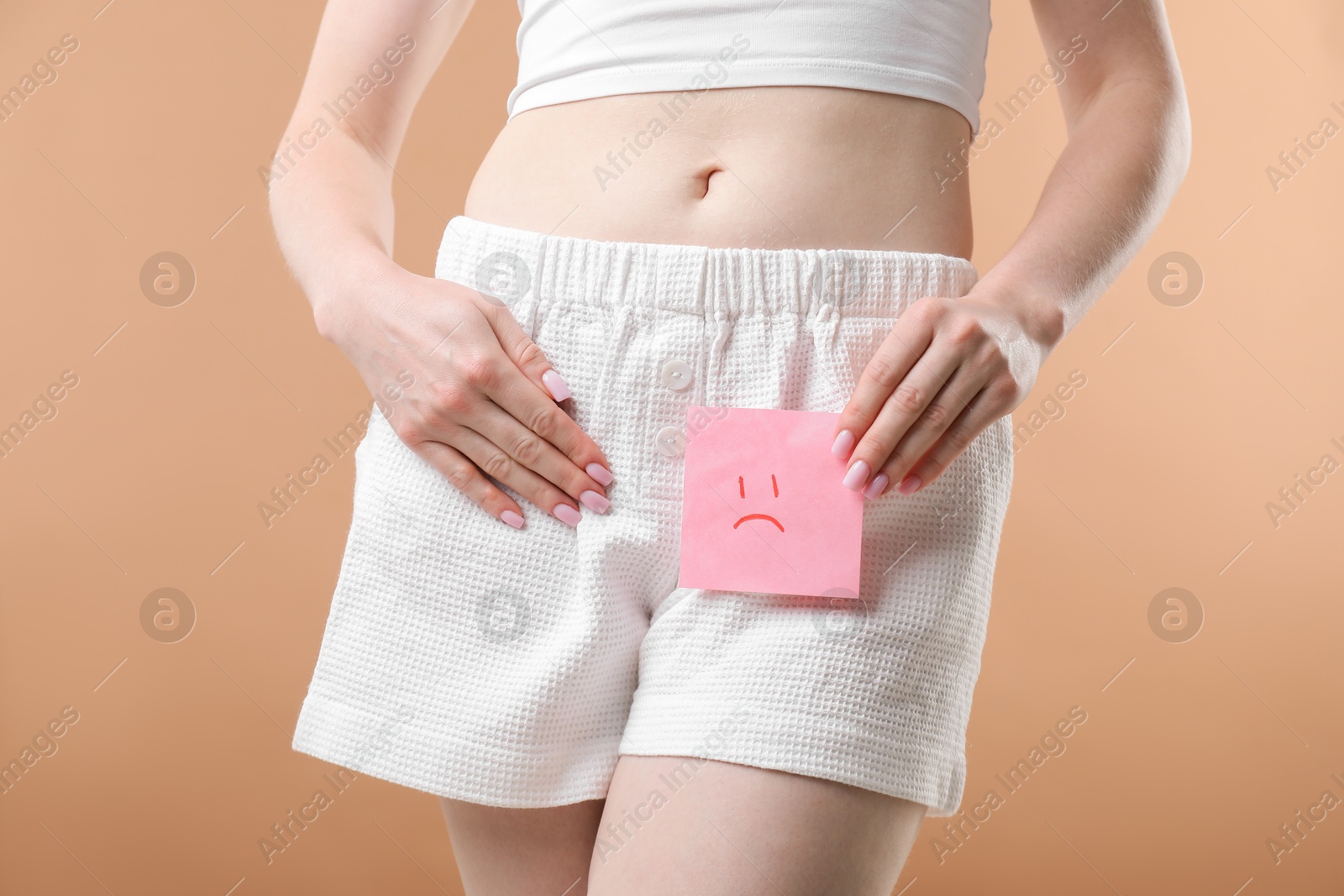 Photo of Woman holding sticky note with drawn sad face and suffering from cystitis symptoms on dark beige background, closeup