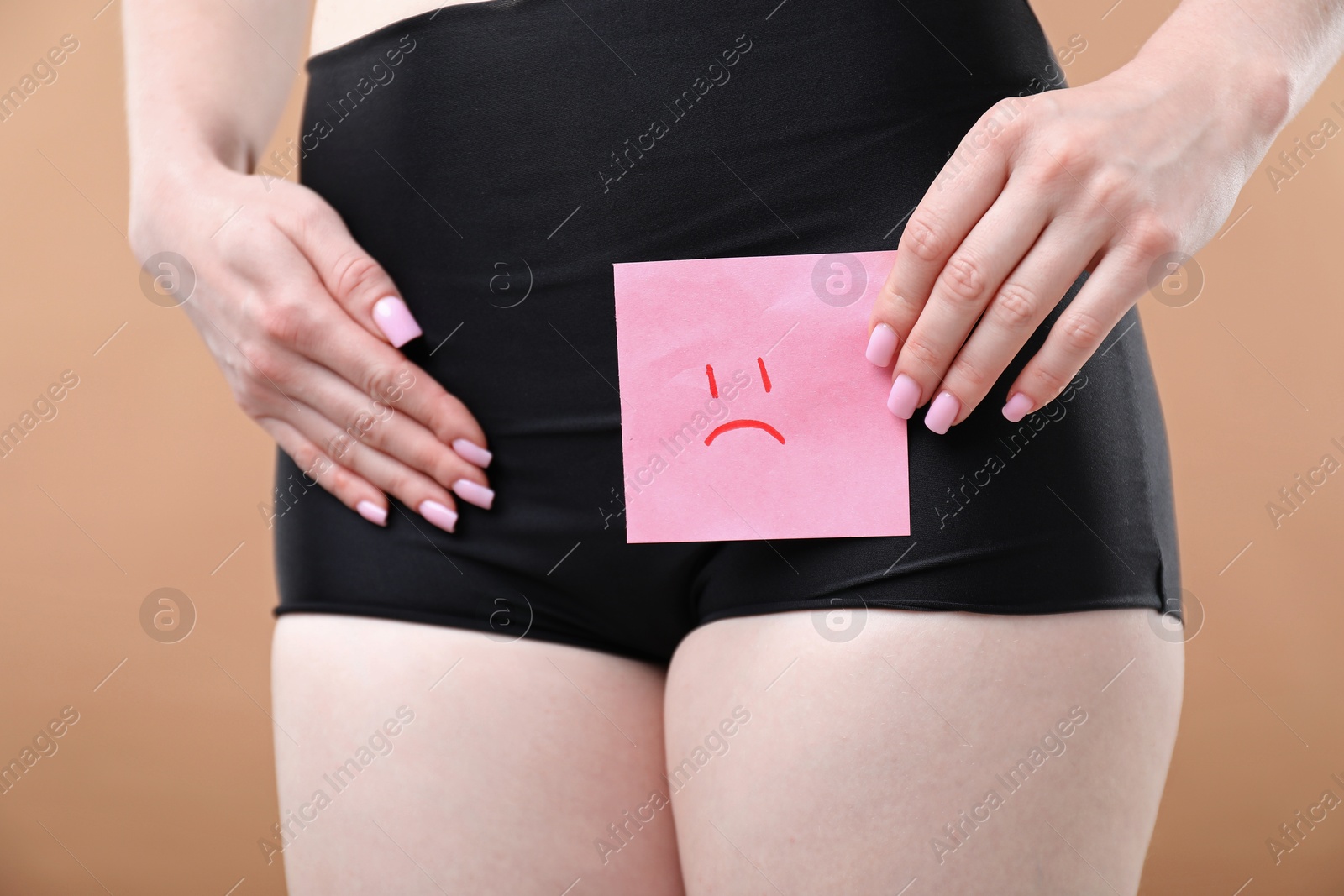 Photo of Woman holding sticky note with drawn sad face and suffering from cystitis symptoms on dark beige background, closeup