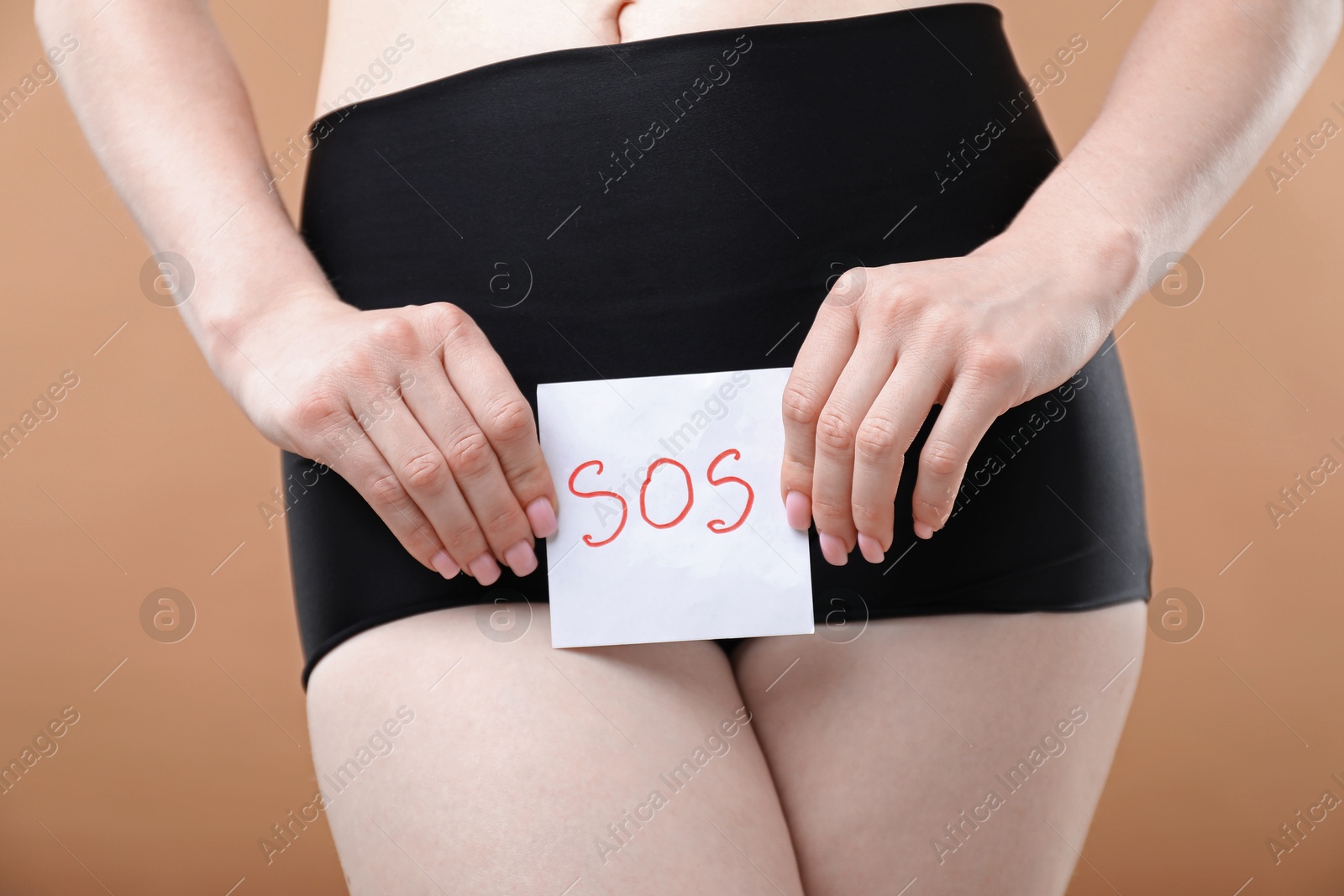 Photo of Woman holding sticky note with word SOS and suffering from cystitis symptoms on dark beige background, closeup