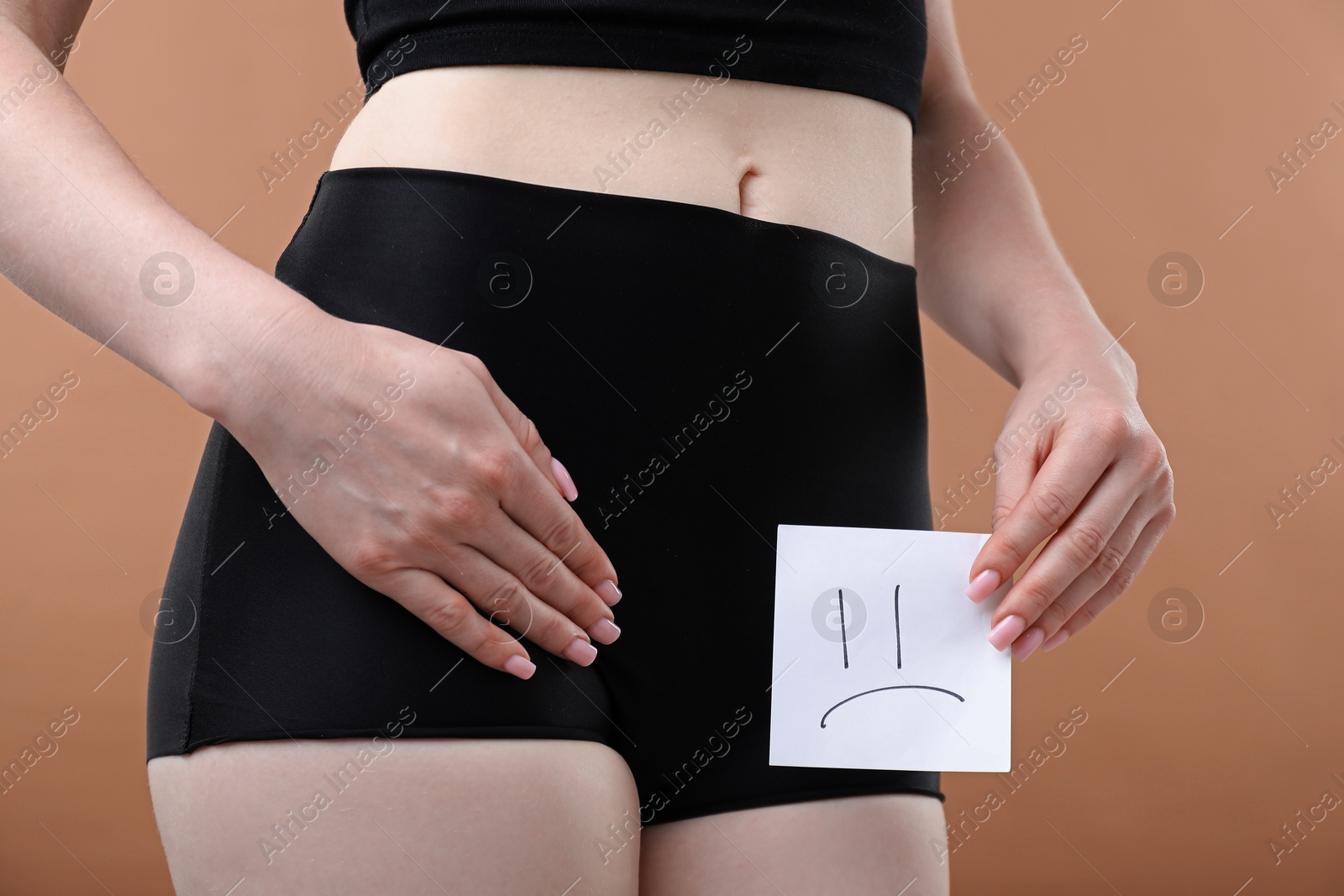 Photo of Woman holding sticky note with drawn sad face and suffering from cystitis symptoms on dark beige background, closeup