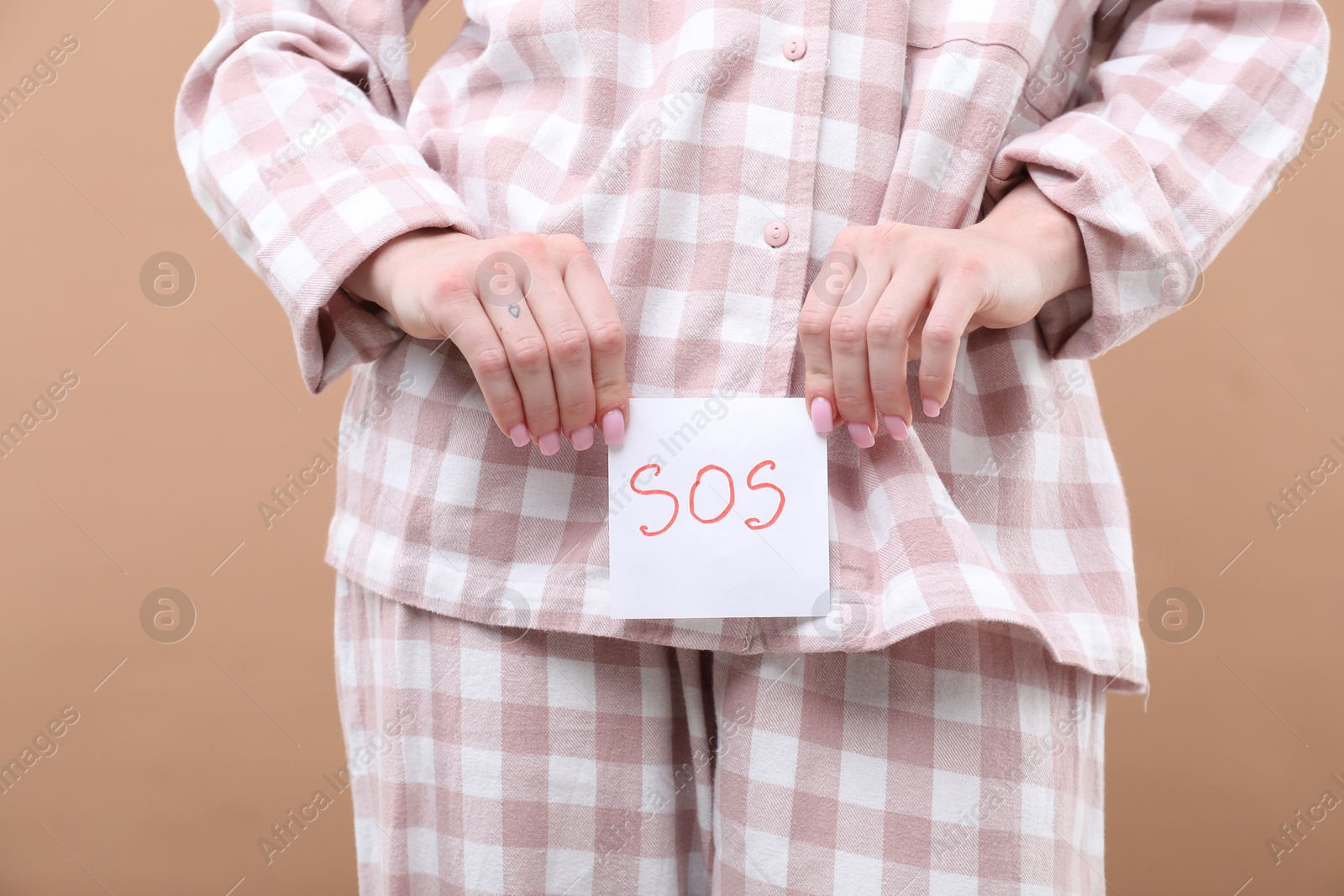 Photo of Woman holding sticky note with word SOS and suffering from cystitis symptoms on dark beige background, closeup